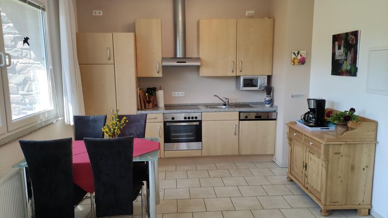 Eine helle, moderne Küche mit Holzschränken und einem Esstisch für vier Personen. Im Hintergrund sind Küchengeräte und eine Fensterfront zu sehen.