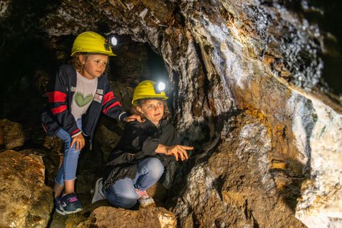 Zwei Kinder erkunden eine Höhle und tragen Schutzhelme mit Lampen. Sie blicken neugierig auf die Felsen und das Umfeld.