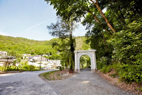 Weißes Tor mit der Aufschrift ‚Römerkessel – Das Leben erwartet dich‘ steht am Beginn eines Spazierwegs im Grünen. Der Weg führt durch einen von Bäumen gesäumten Park. Links im Hintergrund liegen Häuser eines kleinen Ortes vor bewaldeten Hügeln unter blauem Himmel.