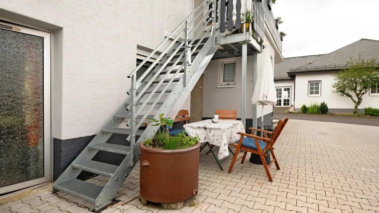 Eine Treppe führt zu einem Eingang, umgeben von einem gepflasterten Hof. Zwischen der Treppe und einem Tisch mit Stühlen steht ein großer Blumentopf.