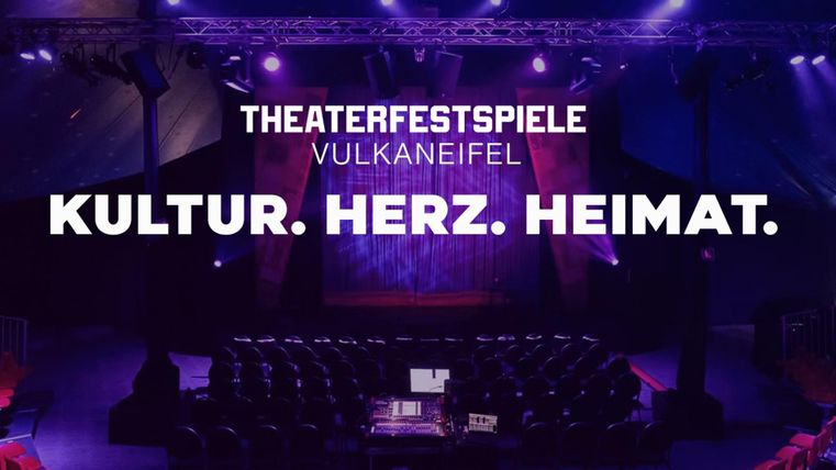 Ein Veranstaltungssaal mit roten Stühlen und einer Bühne im Hintergrund. Die Beleuchtung ist in Violett gehalten und vermittelt eine einladende Atmosphäre.