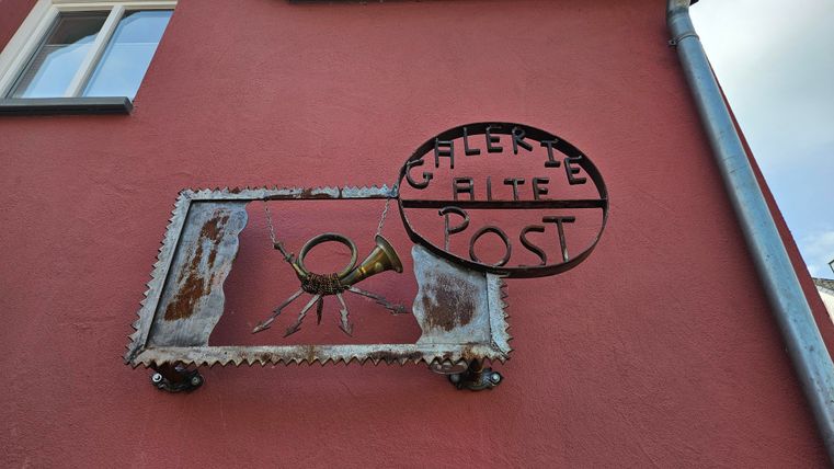 Ein großes Schild mit der Aufschrift "Galerie Alte Post" und einer Kranichgrafik ist an einer rosa Wand angebracht. Das Schild hat einen rustikalen Stil und zieht die Aufmerksamkeit auf sich.