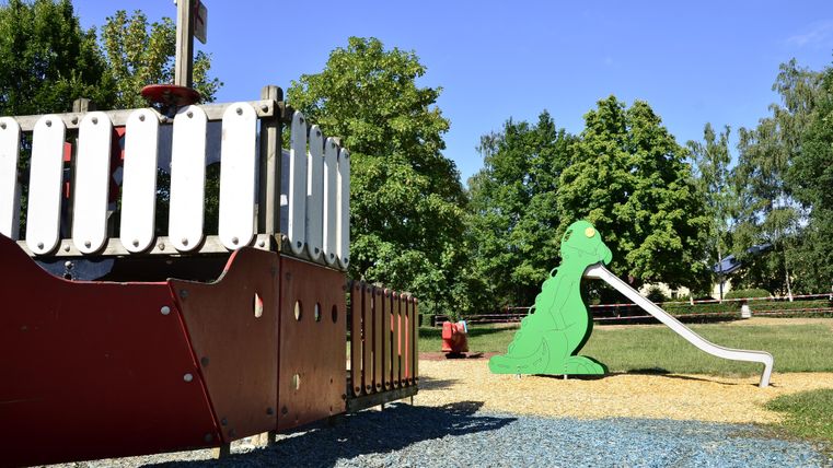 Een speeltuin met een rood speelschip en een groene glijbaan in de vorm van een dier. Op de achtergrond zijn bomen en een heldere blauwe lucht te zien.