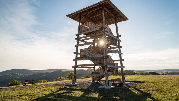Ein hölzerner Aussichtsturm auf einer Wiese mit Blick auf eine hügelige Landschaft bei Sonnenuntergang.