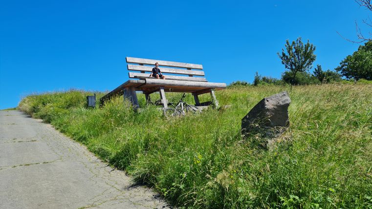 Un grand banc en bois sur une pelouse avec une personne dessus, entourée d'herbe verte et d'un ciel bleu clair.