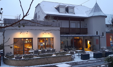 Ein einladendes Weinrestaurant mit hübscher Weinrebe im Vordergrund. Im Hintergrund sieht man das Restaurant und eine schöne Landschaft mit Weinbergen.