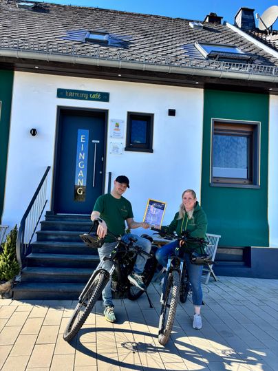 Twee personen staan met fietsen voor een gebouw met de tekst „harmony café“. Het is een zonnige dag en de sfeer lijkt uitnodigend.