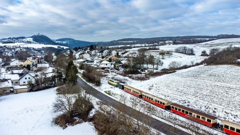 A colorful locomotive stands in the snow while people stroll on the platform. The landscape is wintry and picturesque with snow-covered trees.