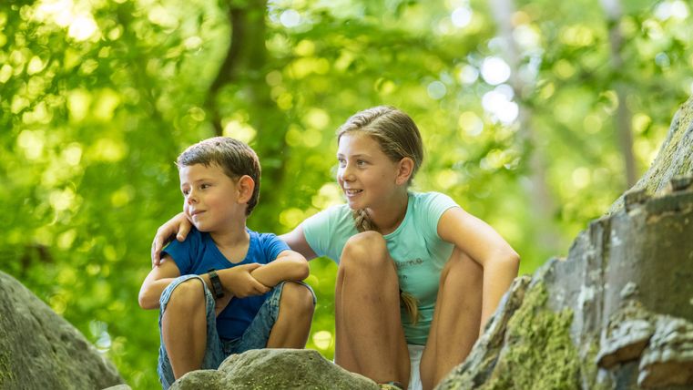 Twee kinderen zitten op rotsen in het bos, omringd door groen gebladerte.
