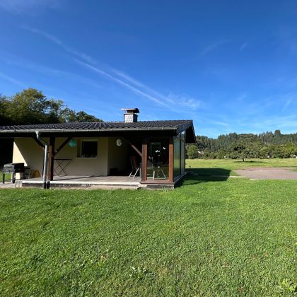 Une maison simple sur une prairie avec de l'herbe verte. En arrière-plan, on voit des arbres et un ciel clair et bleu.
