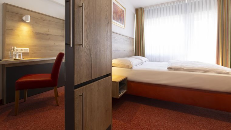 A modern hotel room with a double bed and a desk. The room has large windows and a friendly decor.