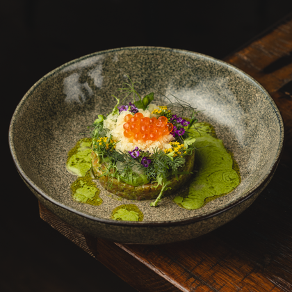 An artistically arranged plate with fresh herbs, colorful edible flowers, and orange fish roe. The background is dark, and the plate is set on a rustic wooden table.