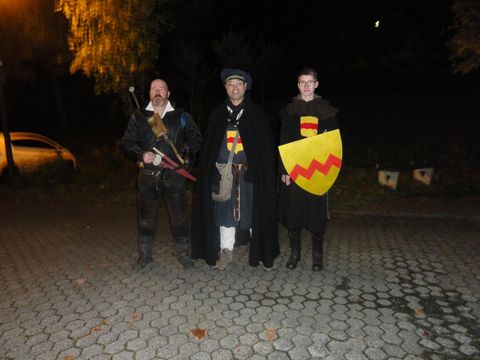 Three men in historical costumes stand in a paved square at night. One holds a sword while another carries a shield.