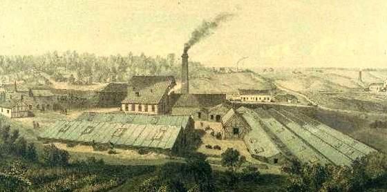 Eine historische Fabrikansicht mit mehreren Gebäuden und einem schornstein, aus dem Rauch aufsteigt. Die Umgebung ist ländlich mit Bäumen und Feldern.