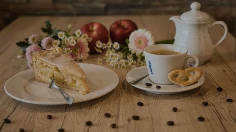 Een stuk appeltaart op een bord naast een kop thee. Op de achtergrond zijn appels, bloemen en koffiebessen te zien.