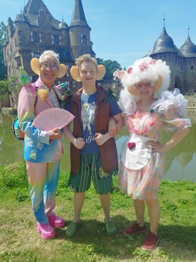 Drie mensen in kleurrijke kostuums staan voor een kasteel. Ze glimlachen en lijken veel plezier te hebben.