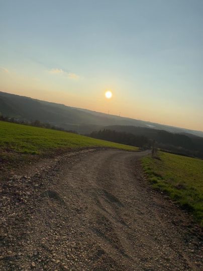 Ein Weg führt durch eine grüne Landschaft, mit einem Sonnenuntergang im Hintergrund. Der Himmel ist klar und die Stimmung ist friedlich.