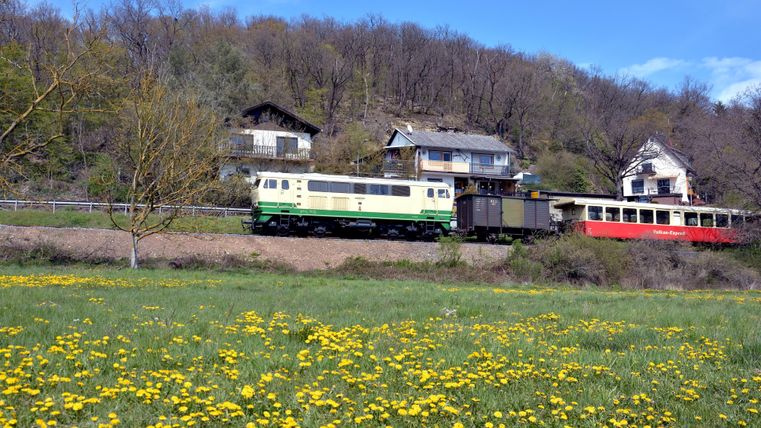 A green locomotive stands on a snowy railway track. In the background, snow-covered trees and a red wagon can be seen.