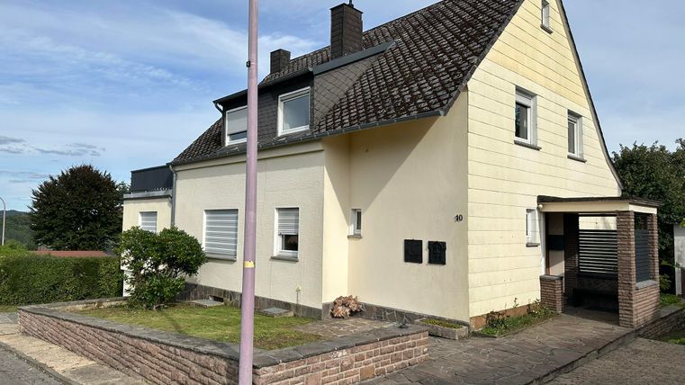 Une maison lumineuse à deux étages avec un toit en pente. Elle est située dans une rue tranquille, entourée d'espaces verts.