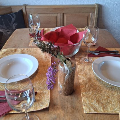 Een mooi gedekte tafel met twee borden, glazen en servetten. In het midden staat een schaal met rode servetten en een kleine vaas met bloemen.