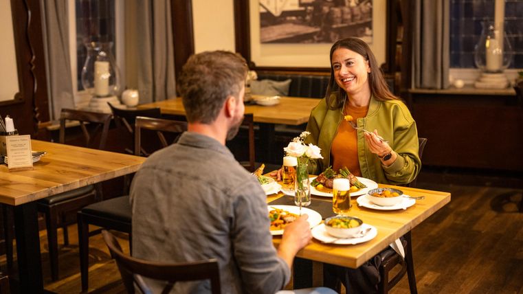 Ein Paar sitzt lächelnd an einem Tisch in einem Restaurant. Auf dem Tisch sind zwei Teller mit Essen und Gläser mit Bier.