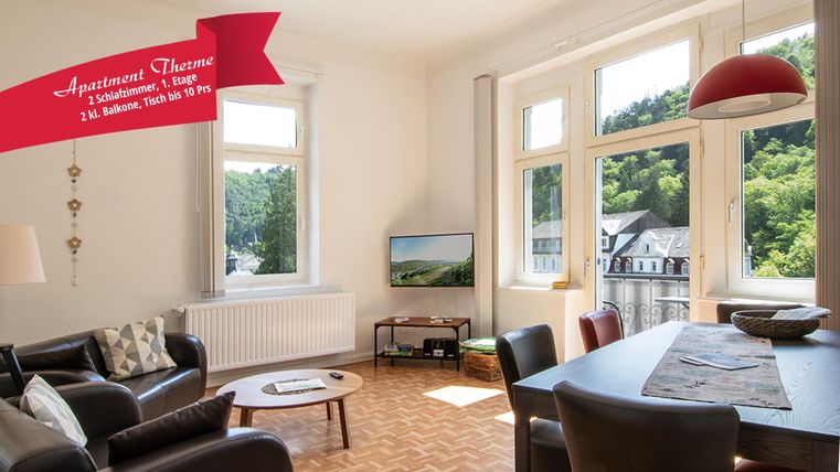 Un salon moderne avec de grandes fenêtres donnant sur un paysage verdoyant. La pièce est lumineuse et accueillante, avec un canapé confortable et une table à manger.