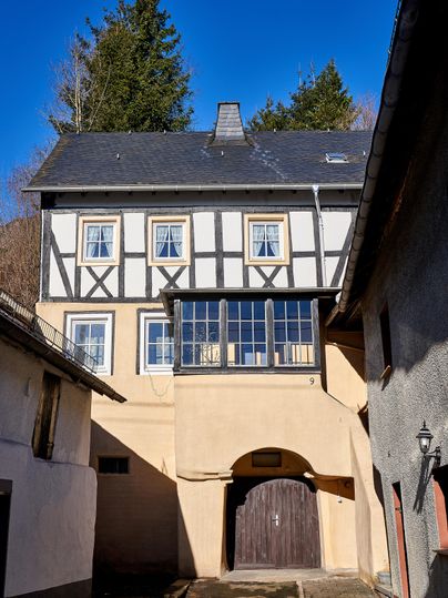 Ein historisches Gebäude mit Fachwerk und einem großen Fenster im Obergeschoss. Der klare blaue Himmel und die umgebenden Bäume verleihen der Szene eine ruhige Atmosphäre.