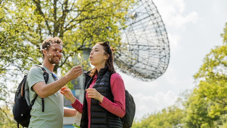 Two people blowing dandelions in front of a large outdoor radio telescope.