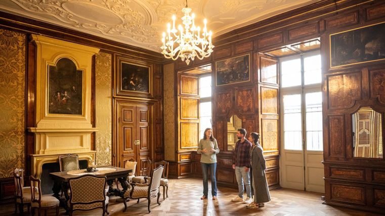 Une chambre élégante avec des murs en bois, décorée avec des peintures. Trois personnes se tiennent autour d'une table et discutent.