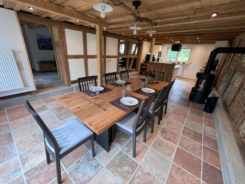 A cozy dining area with a large wooden table and chairs. The room has wooden beams and a modern kitchen in the background.