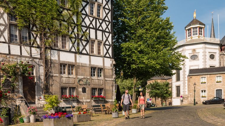 Deux personnes se promènent dans une rue pavée de Kornelimünster, devant une maison à colombages et un café.