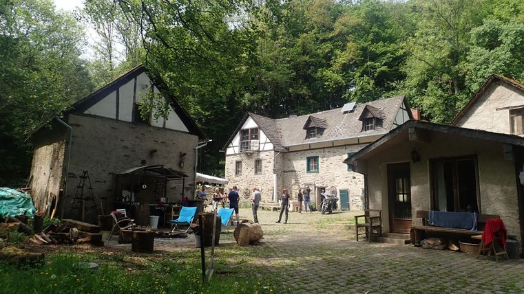 Eine malerische Anlage mit mehreren Gebäuden inmitten von Bäumen. Der Hof ist mit Sitzgelegenheiten und Werkzeugen ausgestattet, was auf eine kreative Atmosphäre hinweist.