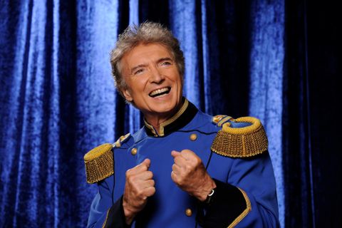 Un homme souriant en uniforme bleue avec des épaulettes dorées. L'arrière-plan est bleu foncé et crée une atmosphère festive.