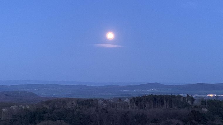 Der Mond leuchtet am Abendhimmel über einer hügeligen Landschaft. Die Umgebung ist düster und ruhig, mit Bäumen im Vordergrund.