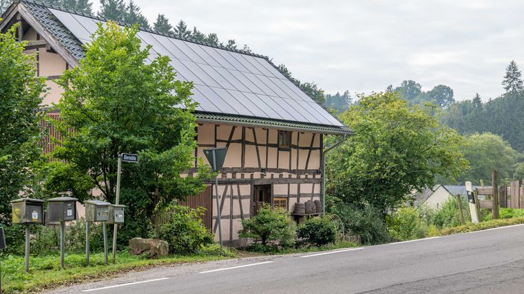 Fachwerkhaus mit Solarpanelen auf dem Dach, umgeben von Bäumen, an einer Straße mit Briefkästen.