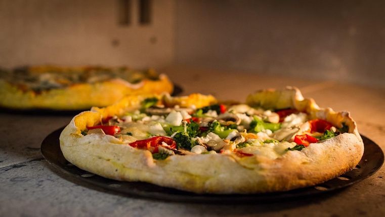 Zwei mit reichlich Gemüse belegte Pizzen liegen auf jeweils einem runden Blech in einem Steinofen und backen.