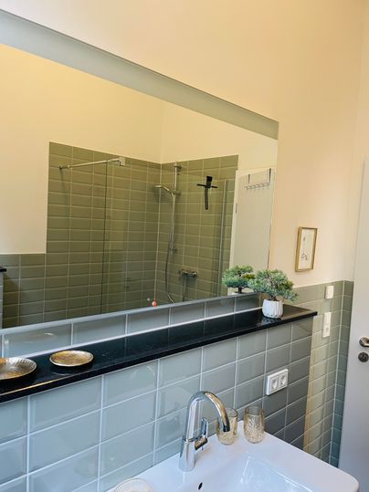 Une salle de bain moderne avec une douche élégante et une surface de miroir claire. Les murs sont carrelés dans des teintes vertes douces, et il y a des éléments décoratifs sur le lavabo.