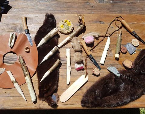 A variety of handcrafted tools and materials lie on a table. Among them are wooden pieces, stone tools, and a piece of leather.
