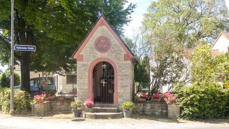 Kleine stenen kapel met bloemen en bomen op de achtergrond.