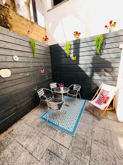 Een gezellige binnenplaats met grijze muren en een blauwe tapijt. Er is een ronde tafel met stoelen en enkele bloemen aan de muur.