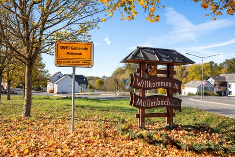 Ein Willkommensschild in der Gemeinde Müllenbach steht an einer Straße. Im Hintergrund sind einige Häuser und herbstliche Bäume zu sehen.