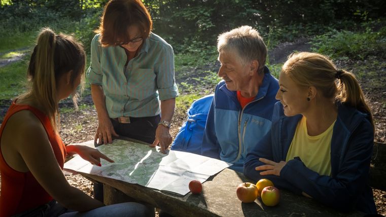 Vier mensen zitten aan een houten tafel in het bos en bekijken een kaart. Er liggen appels op tafel.