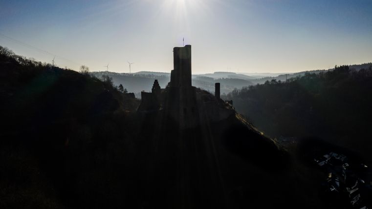 Eine imposante Burg auf einem Hügel, umgeben von sanften Hügeln und der Morgenstrahlung. Der Himmel ist klar und strahlend.