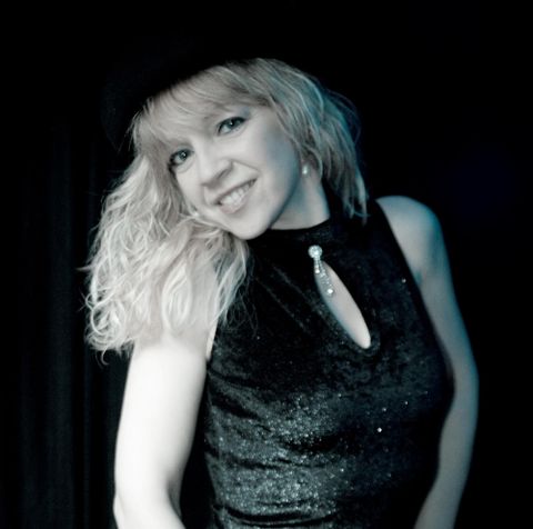 Een glimlachende vrouw met blond, krullend haar draagt een zwart, glinsterend jurk. De achtergrond is donker, wat de focus op haar legt.