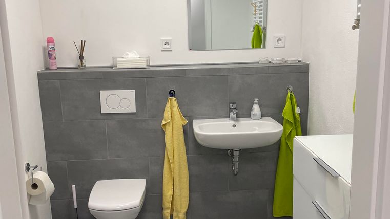 Ein modernes Badezimmer mit grauen Fliesen und einer weißen Toilette. An der Wand hängen gelbe und grüne Handtücher neben einem Waschbecken.