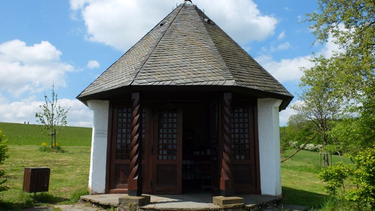 Ein achteckiger Pavillon mit Holztüren und einem Schindeldach in einer grünen Landschaft.