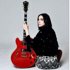 A woman is sitting on the floor holding a red guitar. She is wearing a black sweater and has long, dark hair.