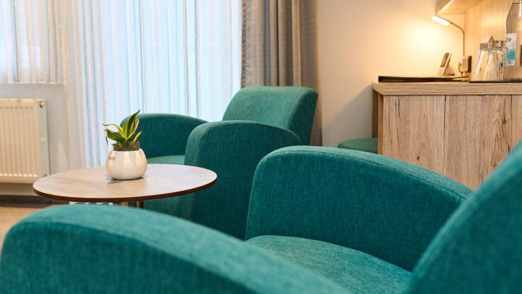 Twee groene fauteuils staan naast een kleine tafel met een plant. Op de achtergrond is een lichte kamer te zien met een houten sideboard.