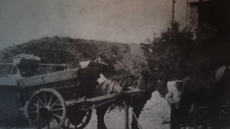 A horse-drawn cart stands still on a street. The wagon is loaded with various goods and pulled by horses.