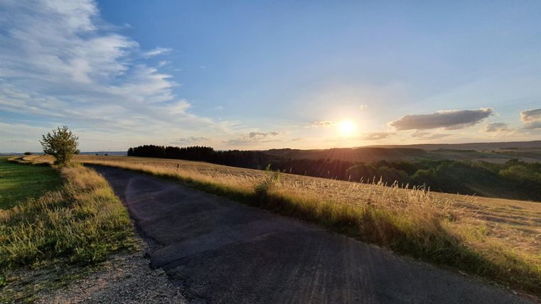 Een schilderachtige weg leidt door open velden onder een heldere lucht. De zon gaat onder aan de horizon en dompelt het landschap in warm licht.